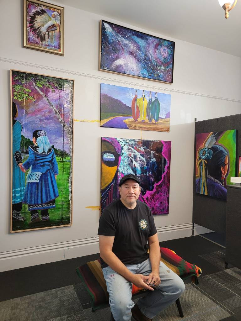 From Blue Bear Studio Facebook photos, Christopher Sweet sitting in front of several of his paintings.